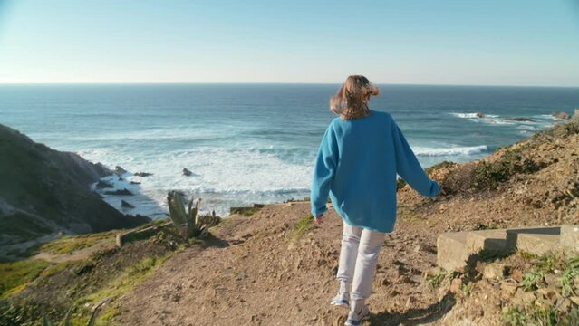 Cinematic adventure concept, young free woman reach end of trail or road on the coastline windy beach. Woman look back at camera after reaching destination on sunny travel day