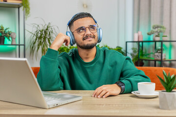 Happy relaxed overjoyed Indian man in wireless headphones dancing at home office table workplace choosing listening favorite energetic disco rock n roll music entertaining. People weekend activities