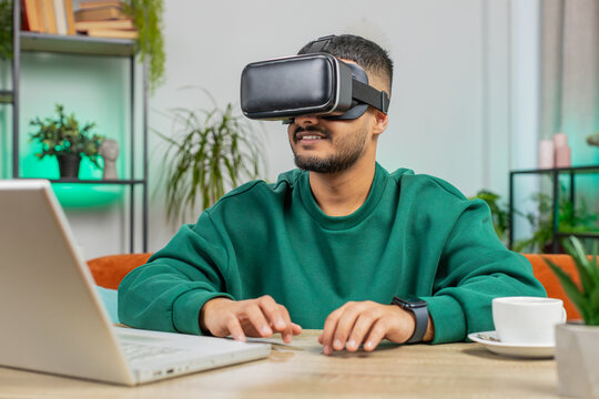 Indian man using virtual reality futuristic technology VR app headset helmet typing on virtual laptop keyboard, play simulation 3D 360 online video game at home office. Guy in goggles sitting at table