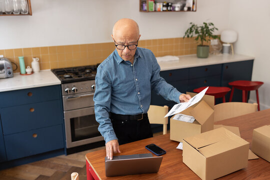 Senior male businessman packing orders into cardboard boxes to ship and using a laptop