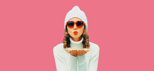 Winter portrait of young woman blowing her lips sending air kiss wearing white knitted sweater and hat, red heart shaped sunglasses on pink studio background