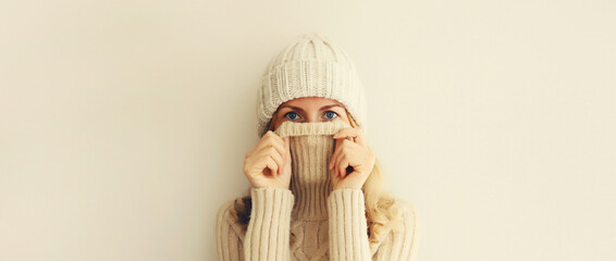 Winter portrait of woman freezing trying to warm up wearing warm soft knitted clothes, hat and sweater on beige studio background