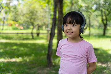 Portrait of child girl listens to music with modern headphones in park outdoors. Happy child enjoying rhythms in listening to music with headphones wireless