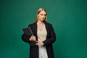 trendy blonde woman in black winter coat holding laptop and looking away on green backdrop
