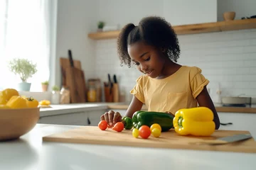 Zelfklevend Fotobehang black African-American girl 10-12 years old in a yellow T-shirt in a light modern kitchen cooks, washes vegetables © Michael