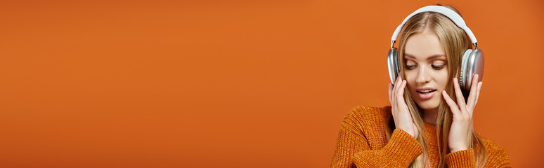 blonde woman in orange knitted sweater and headphones listening music on bright backdrop, banner