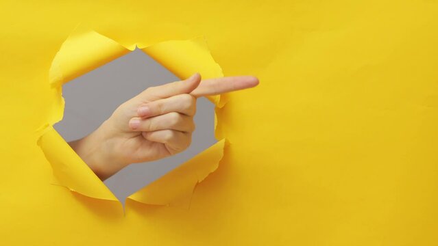 Unrecognizable female hand pointing aside indicating at advertisement area breaking trough paper hole torn on yellow copy space background.