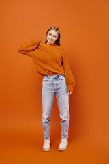 full length of trendy blonde woman in jeans and soft knitted sweater posing on orange backdrop