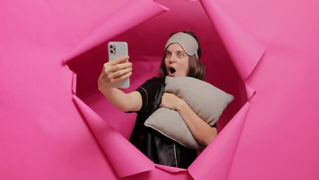 Pleased positive woman wearing sleeping mask and pajama posing in paper hole of pink wall with pillow in hands taking selfie after waking up creating morning content for social network.