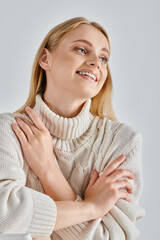 carefree woman with blonde hair sitting in cozy winter sweater and looking away on grey, happiness