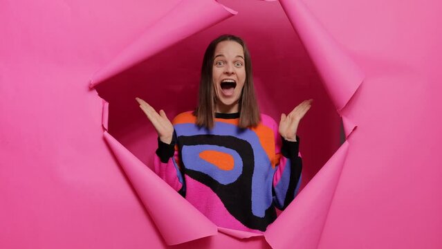 Overjoyed brown haired adult woman wearing stylish jumper standing through torn hole of pink background rejoicing good news sees something excited and amazing expressing happiness.