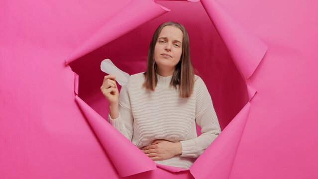 Gloomy Caucasian woman cares about personal hygiene, holding sanitary pad in hands, suffering from pain during menstruation having sad expression breaking through pink paper wall.