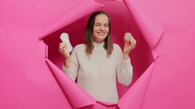 Confused woman holding menstrual cup and hygiene napkin choosing hygiene items for periods breaking through pink paper hole making choice of period protection.