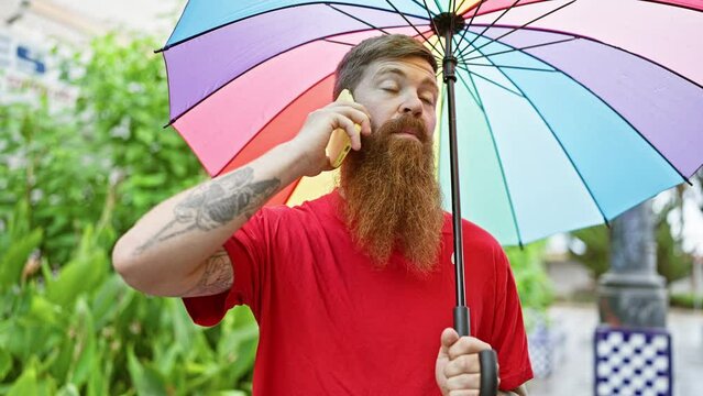 Happy redhead man enjoys a joyful outdoor talk on his smartphone under a rainbow in the park, exuding confident positivity while holding an umbrella, smiling genuinely