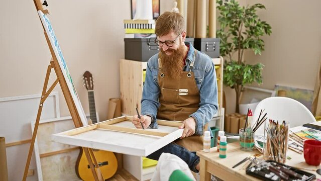 Confident young redhead man rocking the art studio, smiling as he lets his creativity run wild, brush in hand, drawing masterpieces