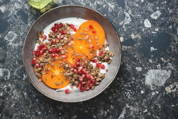 Plate of greek yogurt with granola, sliced persimmon and pomegranate, top view on a dark-brown...
