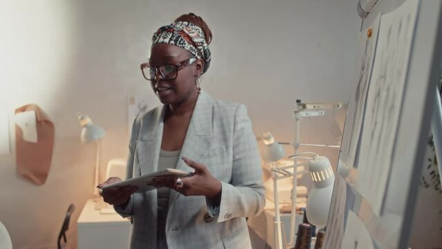 Medium side shot of female African American fashion designer in glasses and suit introducing ideas on clothing collection pointing to sketches on whiteboard and using tablet during meeting in atelier