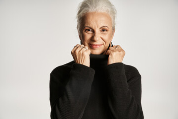 portrait of middle aged business woman adjusting collar of turtleneck sweater on grey backdrop