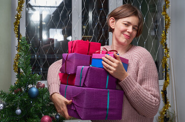 A woman with a pile of Christmas gifts in her hands stands next to a decorated Christmas tree