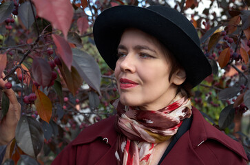 Portrait of a woman in a red cloak and hat against the background of an autumn tree with ripe red berries