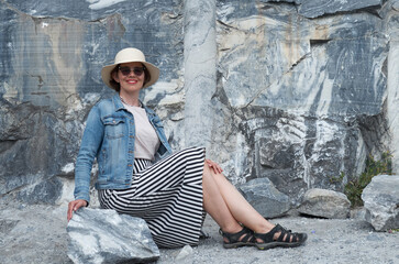 A female tourist in a straw hat and denim jacket poses against the background of a natural marble wall