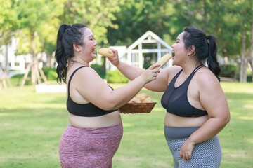 Cheerful two woman overweight in sportswear have fun and enjoy eating bread while exercising in garden. Asian girl chubby holding bread looking at camera standing in the park.
