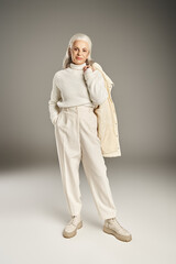 Fototapeta premium happy and grey haired middle aged woman in white outfit holding winter jacket and standing on grey