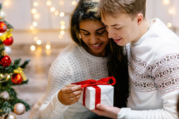 A young couple celebrates Christmas next to a decorated Christmas tree. A man gives his beloved a gift