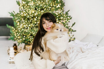 Young pretty woman holding small german spitz dog at her hands and standing against christmas tree with bookeh lights. girl and cute dog celebraiting New Year.