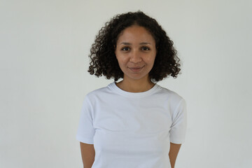 A young woman with curly hair wearing a plain white T-shirt stands against a simple white background, looking directly at the camera with a subtle smile © Kseniya