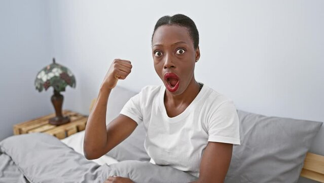 Amazed african american woman in bedroom, lying on bed with a surprised expression, giddily pointing, mouth wide open!
