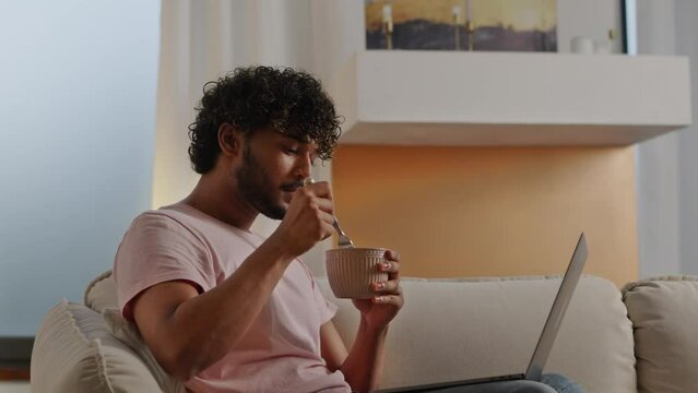 Indian freelancer man eating instant noodles while workg witinh laptop indoors. Young hungry guy sitting on comfortable couch watching movie on computer. Male student studying online and having a