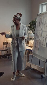 Vertical full side shot of African American female creative director in suit and glasses presenting ideas for new clothing collection standing near flipchart with sketches and using tablet in atelier
