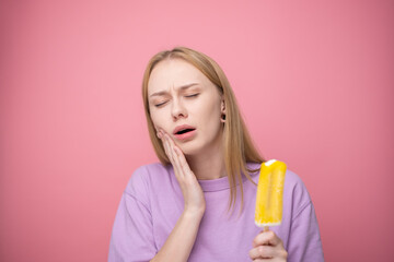 Sad young woman eating yellow ice cream on pink background. Concept of sensitive teeth to cold...
