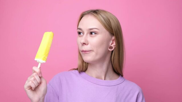 Happy young woman eating yellow ice cream on pink background. Concept of summer and sweet cold desserts.
