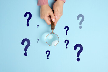 Female hand holds a wooden magnifying glass and question marks on a blue background. The concept of finding an answer to questions - obrazy, fototapety, plakaty