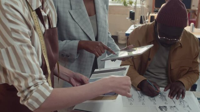 Side tilt shot of atelier colleagues choosing colour for new garment using samples of fabrics and consulting fashion designer who drawing sketches in tailoring atelier