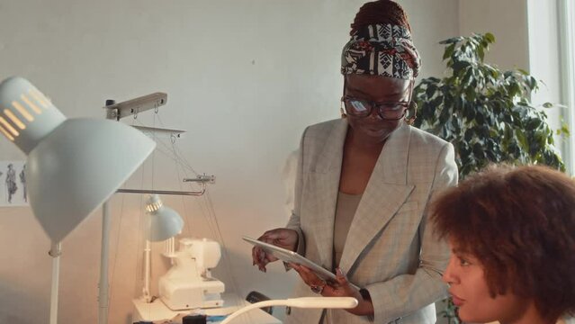 Tilt arc shot of smiling seamstress discussing garment production with female boss who showing design idea on tablet and then going away in tailoring studio