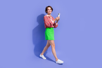 Fototapeta na wymiar Full size photo of good mood lovely woman dressed print shirt holding smartphone walk to empty space isolated on blue color background