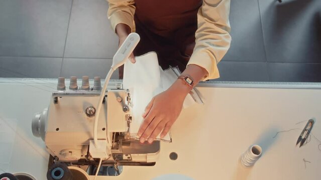 Top view of female dressmaker in apron sitting at table and tailoring white cloth with black seams using sewing machine during workday at atelier