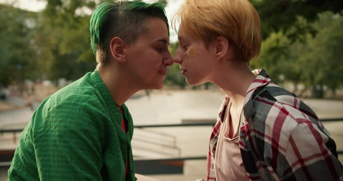 Two lesbian girls, a girl with a green haircut and green hair in a green shirt and a girl with yellow hair in a plaid shirt, lean their faces against each other and rub their noses during their date