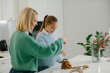 Happy mother and cute girl daughter decorating Christmas gingerbread cookies after baking while standing in cozy kitchen at home with Christmas decorations. - Powered by Adobe