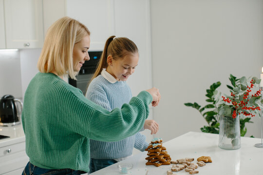 Happy Mother And Cute Girl Daughter Decorating Christmas Gingerbread Cookies After Baking While Standing In Cozy Kitchen At Home With Christmas Decorations.