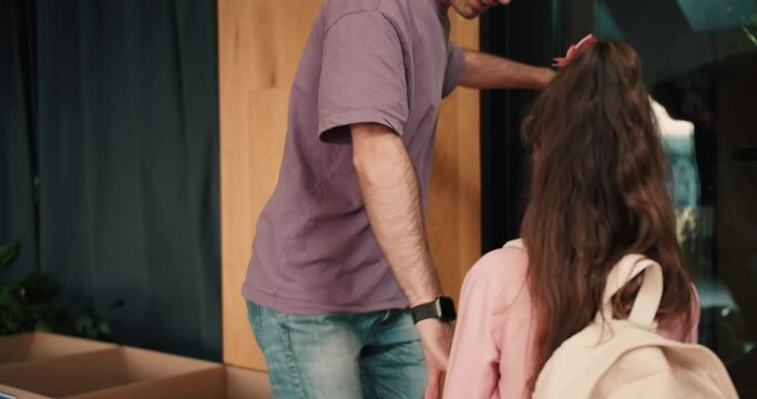 A man with gray hair in a purple T-shirt picks up his daughter in a pink dress and with a white backpack from a group for preparing preschoolers for school