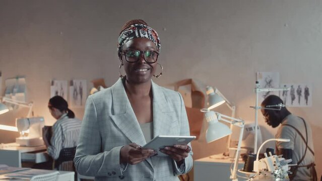 Medium portrait of stylish smiling female African American owner of tailoring studio wearing grey suit and glasses using tablet and then posing looking at camera with working tailors in background