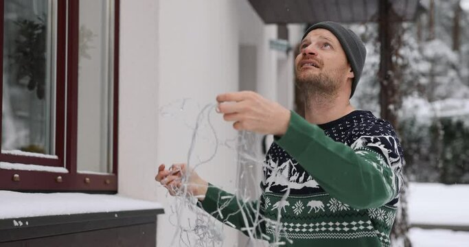 man outdoors in christmas sweater untangling string lights for house exterior decoration