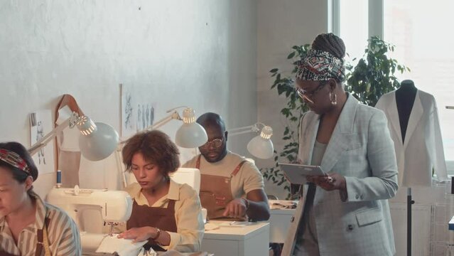Medium pan shot of African American female atelier boss with glasses and tablet walking around and controlling diverse tailors working on sewing machines during garment manufacturing process