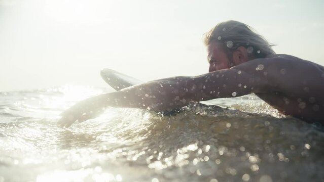 Close-up shot of a blond man with a beard rowing with his hands and splashing water in the sea while surfing. Close-up shot of dynamic swimming of a male surfer with a beard