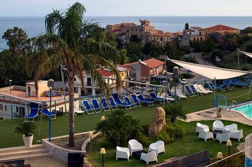 Panoramic landscape view of hotel's swimming pool area with many sun beds. Blue sky over sea in the background. Sunny autumn day. Travel and tourism concept. Tusa, Sicily, Italy