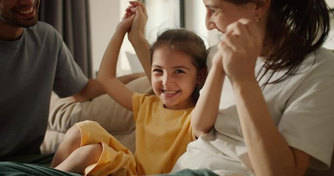 Close Up Shot Of A Brunette Mom In A White T-shirt Playing With Her Daughter In A Yellow Dress While Sitting On A Light Brown Sofa With Her Dad In A Modern Studio Room